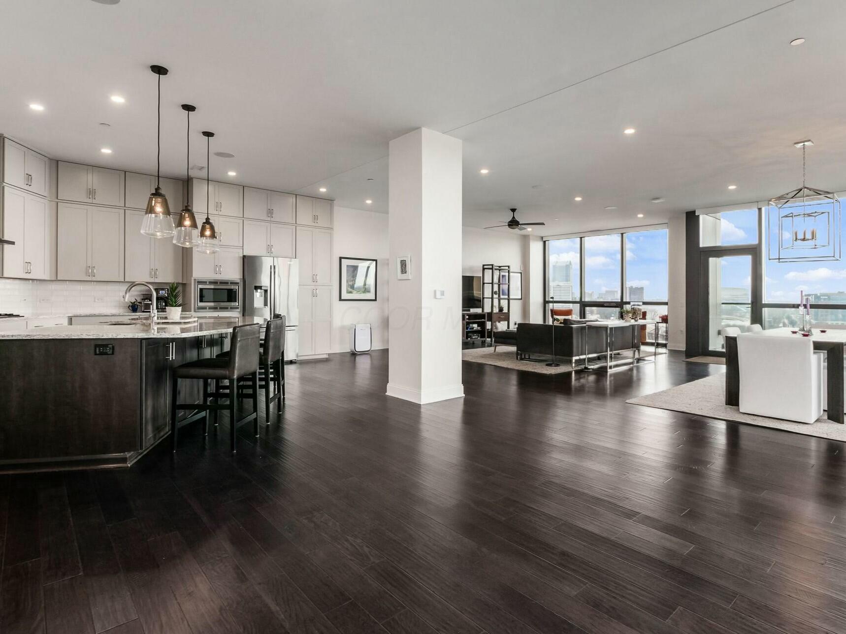 Wide open-plan condominium interior with dark hardwood floors, white kitchen cabinetry, island seating and a wall of windows onto the Columbus skyline