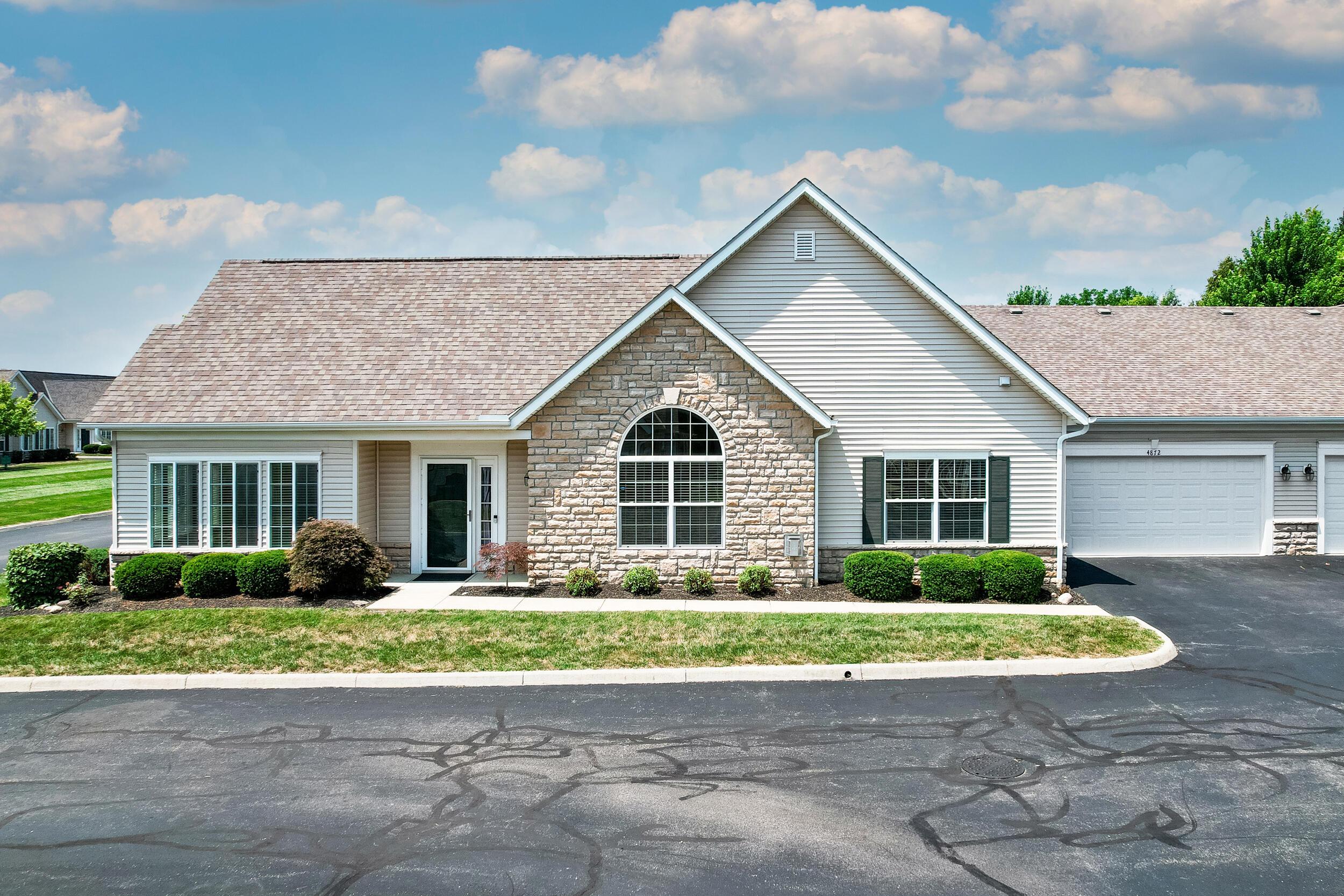 Single-story Dublin condominium with cream siding, a stone-clad gable with an arched window, trimmed shrubs and an attached two-car garage