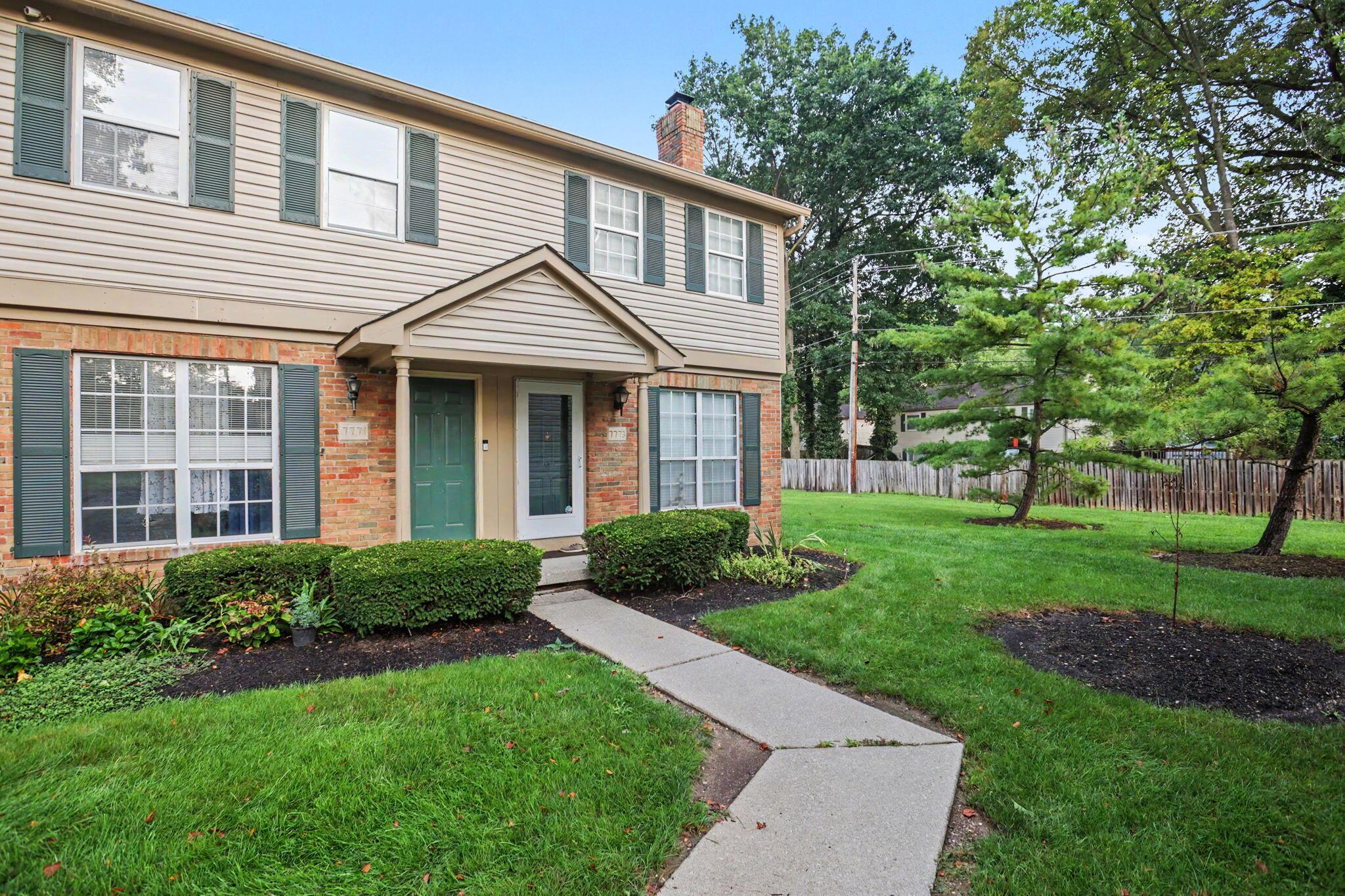 Two-story Dublin townhome with tan siding, brick lower level, green shutters and a covered entry, with a lawn and mature trees alongside