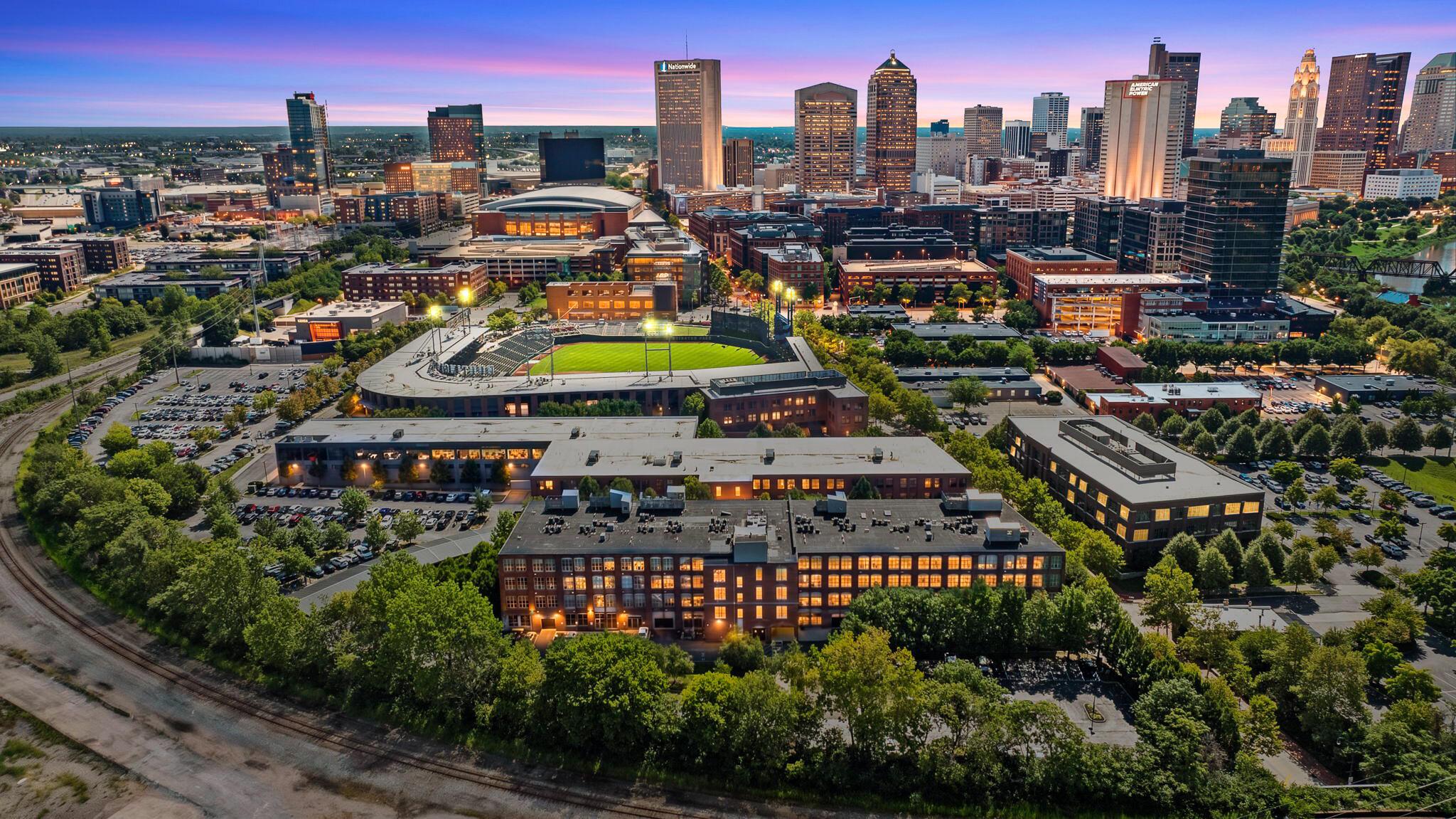 Aerial view of the Arena District in Columbus, Ohio at dusk, with Huntington Park, brick loft buildings and the downtown skyline behind them