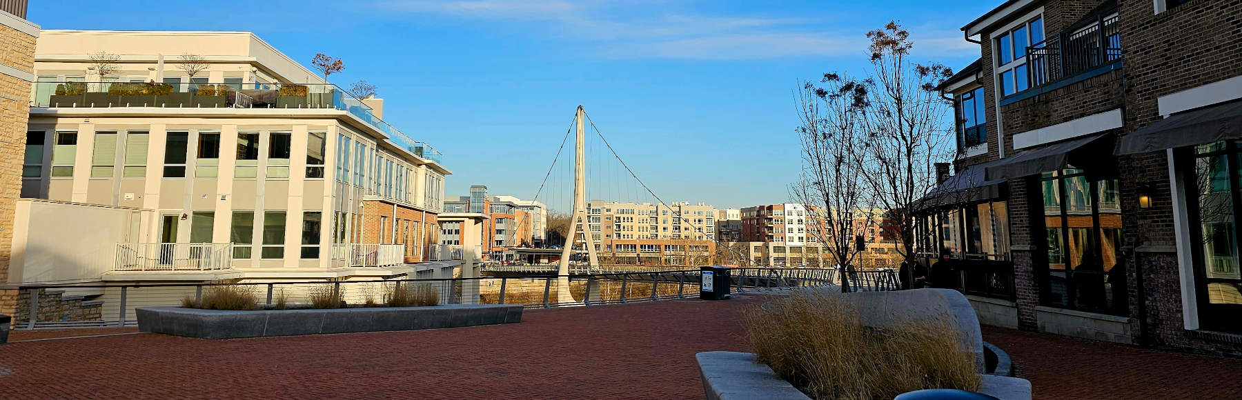 A brick plaza in Bridge Park, Dublin, framed by condominium buildings and restaurant patios, with the tower of the Dublin Link bridge visible at the end of the street