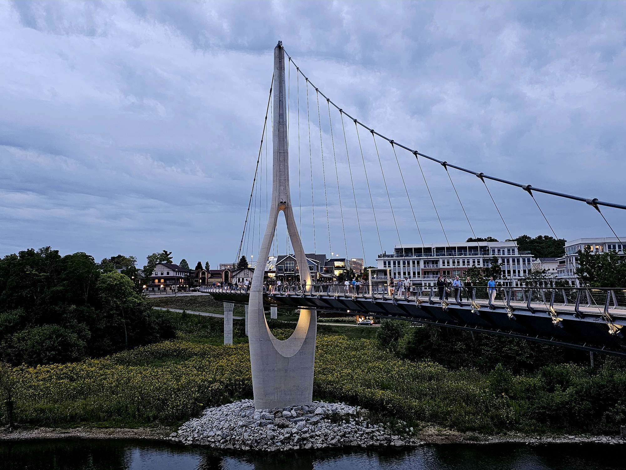 The Dublin Link pedestrian suspension bridge over the Scioto River at dusk, walkers crossing toward the lit restaurants of Historic Dublin and Bridge Park